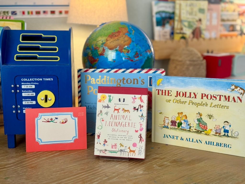 A Mail Monday setup: a toy blue mailbox, a globe, letter-writing picture books, and animal stationery on a wooden table